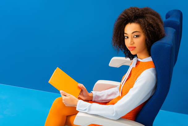high angle view of smiling african american in retro dress holding book on blue background  - Foto, Imagem