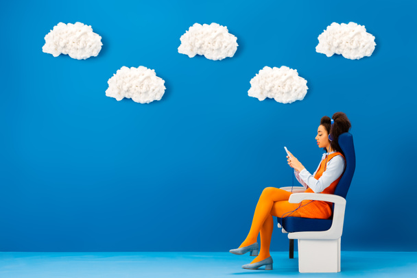 vista lateral de afroamericano en vestido retro escuchando música ti y usando teléfono inteligente sobre fondo azul con nubes
  - Foto, imagen