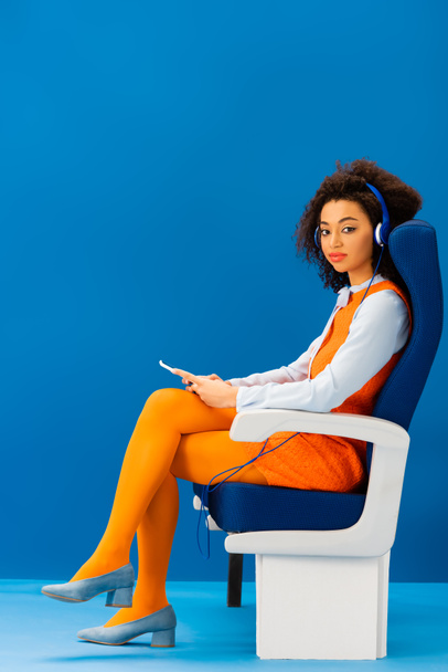 Eski moda elbiseli Afro-Amerikan koltukta oturup mavi arka planda müzik dinliyor.  - Fotoğraf, Görsel