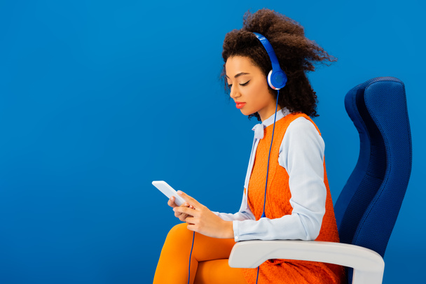 afro-américaine en robe rétro écoutant de la musique et utilisant un smartphone isolé sur bleu
  - Photo, image