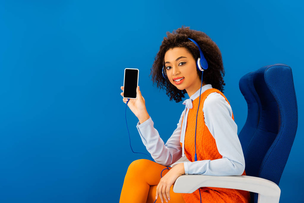 Αφροαμερικάνος με ρετρό φόρεμα ακούγοντας μουσική και κρατώντας smartphone απομονωμένο στο μπλε  - Φωτογραφία, εικόνα