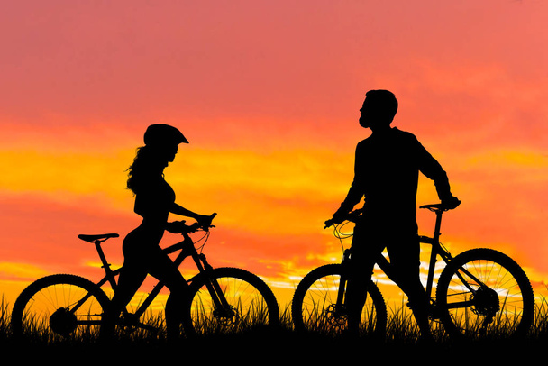 日没の背景に自転車の2つのシルエット。アスレチック男と現代のマウンテンバイクの女の子がお互いに行きます. - 写真・画像