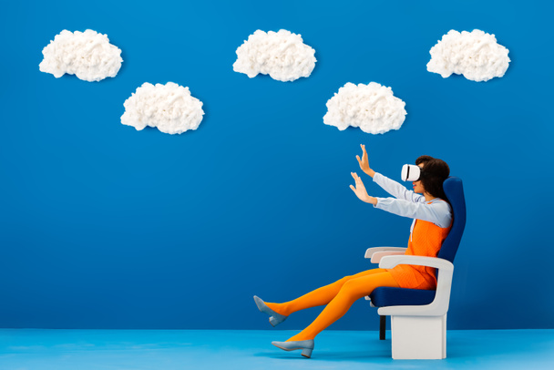 vue latérale du casque afro-américain en vr avec les mains tendues assis sur le siège sur fond bleu avec des nuages
  - Photo, image