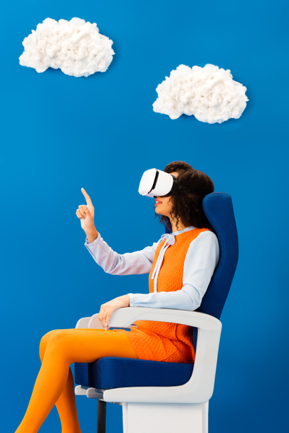 Seitenansicht eines Afroamerikaners mit vr-Headset, das mit dem Finger auf blauem Hintergrund mit Wolken zeigt  - Foto, Bild