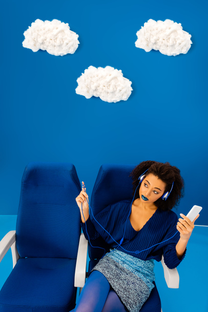 vue grand angle d'afro-américain assis sur le siège, écoutant de la musique et tenant smartphone sur fond bleu avec des nuages
  - Photo, image