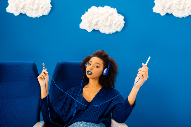 african american sitting on seat, listening to music and using smartphone on blue background with clouds  - Photo, Image