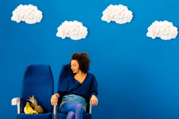 mosolygó afro-amerikai ül az ülésen, és nézi a banán és ananász kék alapon felhők  - Fotó, kép