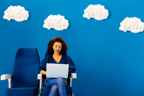 mosolygós afro-amerikai ül az ülésen, és használja laptop kék háttér felhők  - Fotó, kép