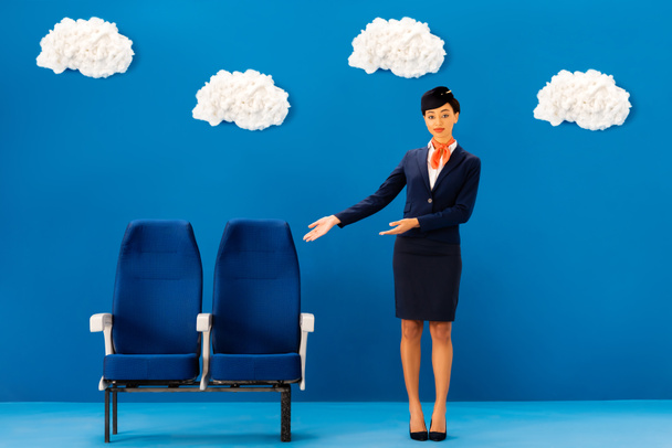 Afrykański amerykański stewardessa wskazując dłońmi na siedzenia na niebieskim tle z chmurami  - Zdjęcie, obraz