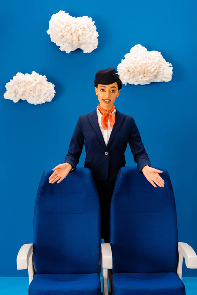 glimlachende Afrikaanse Amerikaanse stewardess wijzend met handen op stoelen op blauwe achtergrond met wolken  - Foto, afbeelding