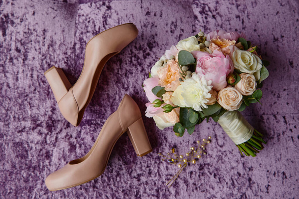 Bridal bouquet of pink peonies, coral roses and greenery with satin ribbon, beige women shoes and hair pins on purple velours armchair, copy space. Wedding concept. Top view, flat lay - Photo, Image