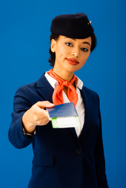 χαμογελαστός Αφροαμερικάνος αεροσυνοδός που κατέχει διαβατήριο και αεροπορικό εισιτήριο απομονωμένο στο μπλε  - Φωτογραφία, εικόνα