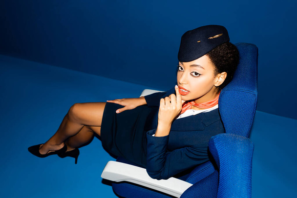 wysoki kąt widzenia afrykańskiej amerykańskiej stewardessy siedzącej na siedzeniu na niebieskim tle  - Zdjęcie, obraz