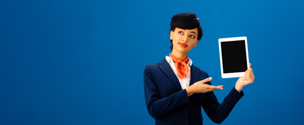 panoramic shot of smiling african american flight attendant pointing with hand at digital tablet isolated on blue  - Foto, immagini
