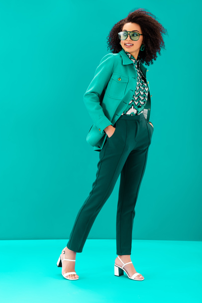 smiling and stylish african american woman looking away on turquoise background  - Photo, Image
