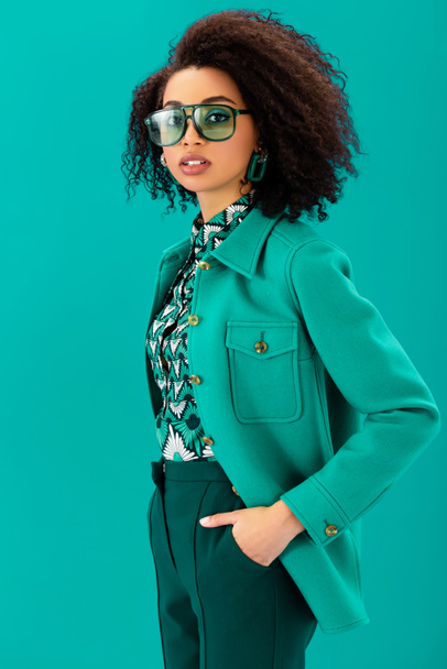 stylish african american woman with hand in pocket looking at camera isolated on turquoise - Foto, Imagem
