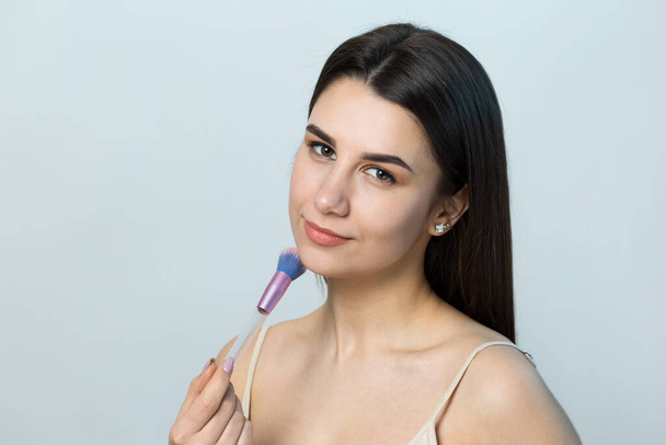 Close-up of a young girl in a light top on a white background making a facial make-up. A pretty woman holds a cosmetic brush near her face and smiles. - Фото, изображение