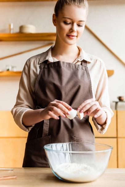 attractive woman in apron adding egg in bowl with flour  - Photo, image