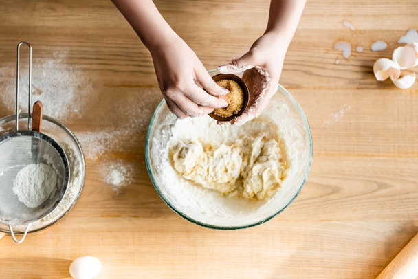 top view of woman holding bowl with brown sugar above dough in bowl, raw eggs and flour  - Foto, Bild