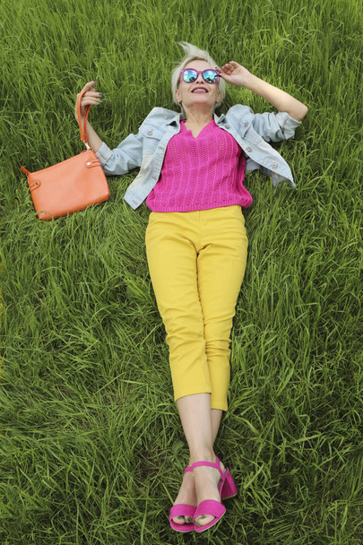 A young woman with a bag is on the background of green grass in yellow pants and a pink jacket with sandals.Urban summer style. - Foto, immagini