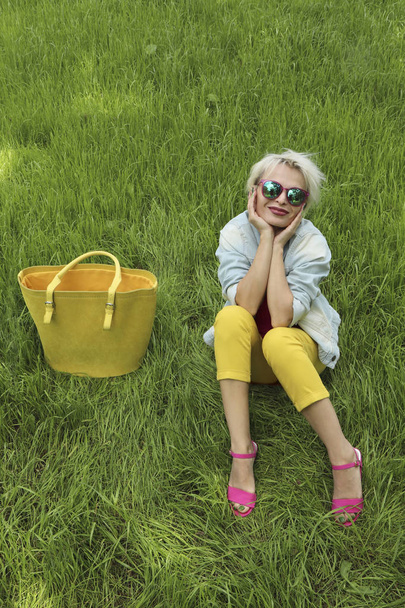 A young woman with a bag is on the background of green grass in yellow pants and a pink jacket with sandals.Urban summer style. - Foto, imagen