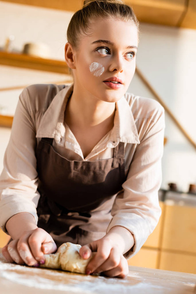 beautiful young woman in apron kneading dough and looking away - Foto, Bild
