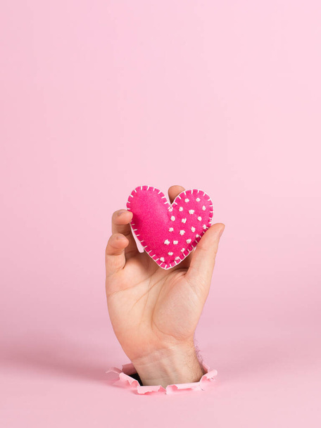 A male hand sticks out of a hole in pink paper, holds a handmade heart. Valentine's day concept, symbol of love, print commercial blank, copy space. - Foto, imagen
