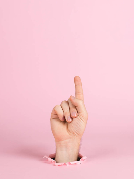 Male hand sticks out of a hole in paper, on a pink background. Indicates a sign, copy space. - Фото, изображение