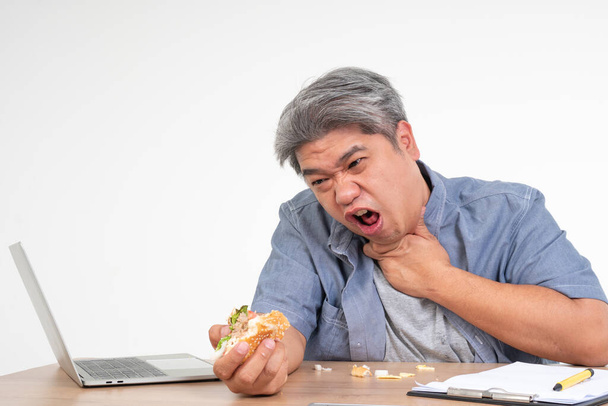 アジア人の男性は、オフィスの机の上でハンバーガーを食べ、食べ物を窒息させた後に首を保持します。忙しいビジネスマンの概念は仕事と健康の世話をしない仕事と左のバランスを取ることができない唯一のジャンクフードを食べる - 写真・画像