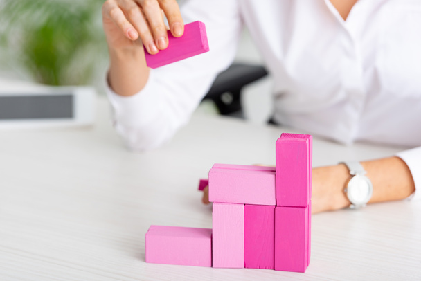 Vista recortada de la mujer de negocios apilando bloques de construcción de color rosa en la mesa
 - Foto, Imagen