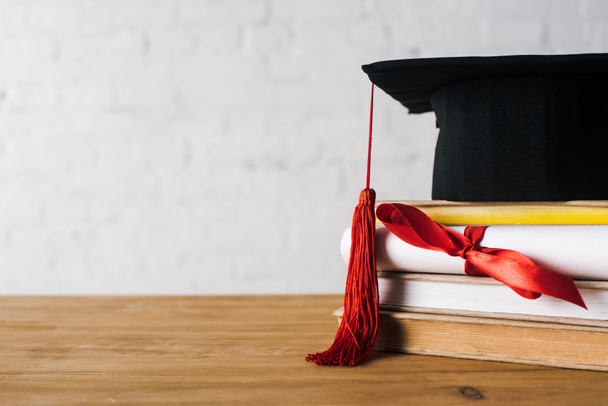 Diploma with beautiful bow, graduation cap with red tassel on top of books on table on white background - Photo, Image