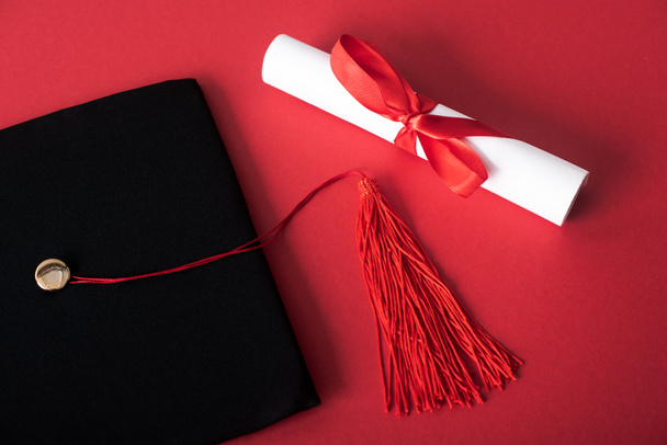 Top view of diploma with beautiful bow and black graduation cap with tassel on red background - Photo, Image