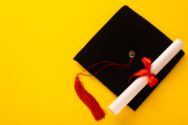 Top view of black graduation cap with red tassel with diploma on top on yellow background - Foto, Bild