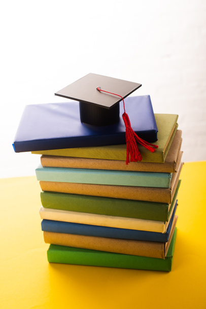 Graduation cap with red tassel on top of books on yellow surface on white background - Photo, Image