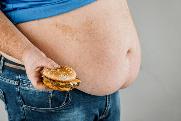 dikke man met een hamburger in zijn hand op een grijze achtergrond. - Foto, afbeelding