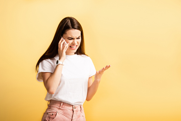 irritated girl standing with open arm and shouting during talk on smartphone isolated on yellow - Photo, Image