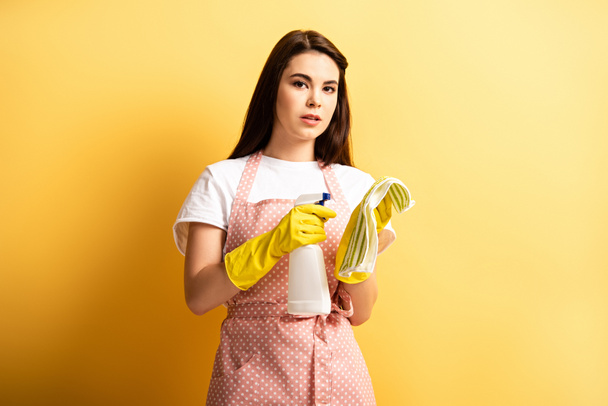 pensive housewife in apron and rubber gloves spraying detergent on rag on yellow background - Foto, Imagen