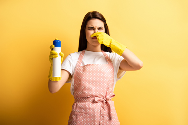 displeased housewife in apron and rubber gloves plugging hose with hand while holding air freshener on yellow background - Photo, Image
