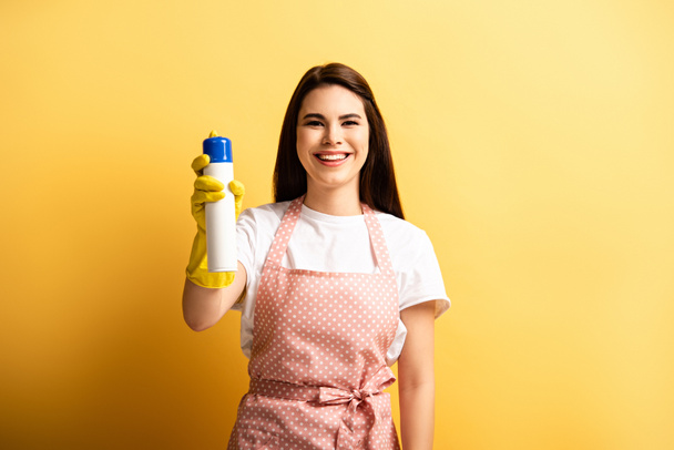 gelukkig huisvrouw in schort en rubber handschoenen kijken naar camera terwijl het houden van luchtverfrisser op gele achtergrond - Foto, afbeelding