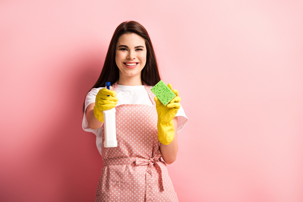 happy housewife in apron and rubber gloves holding spray bottle and sponge on pink background - Zdjęcie, obraz