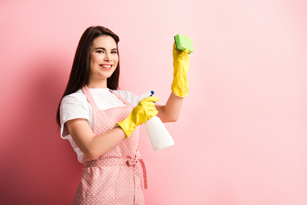 cheerful housewife in apron and rubber gloves holding spray bottle and sponge on pink background - Photo, Image