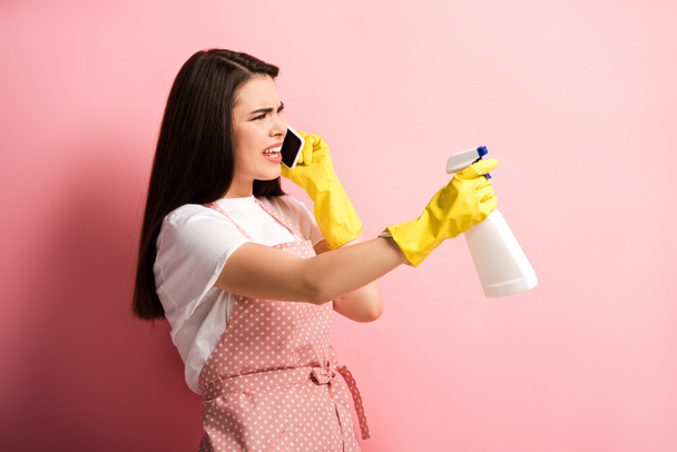 nespokojená žena v domácnosti v zástěře a gumové rukavice mluví na smartphone při držení sprej láhev na růžovém pozadí - Fotografie, Obrázek