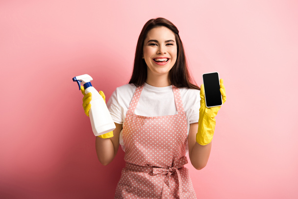 szczęśliwy gospodyni w fartuchu i gumowe rękawiczki pokazując smartfon z pustym ekranem trzymając butelki spray na różowym tle - Zdjęcie, obraz