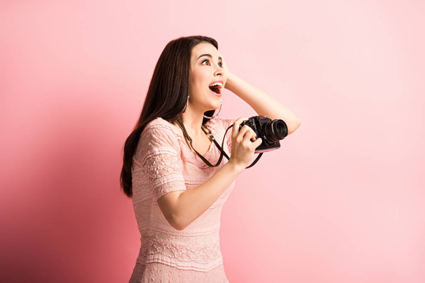 opgewonden fotograaf aanraken hoofd en weg te kijken terwijl het houden van digitale camera op roze achtergrond - Foto, afbeelding