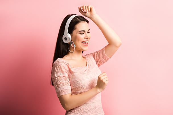 happy girl in wireless headphones dancing and singing on pink background - Photo, Image