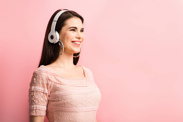 happy girl in wireless headphones looking away while standing on pink background - Photo, Image