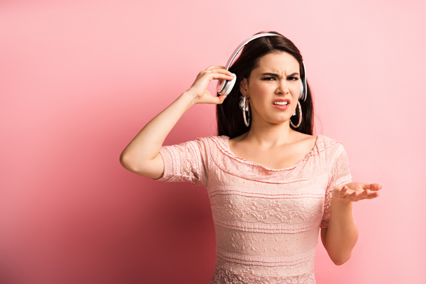 δυσαρεστημένο κορίτσι αγγίζοντας ασύρματα ακουστικά, ενώ στέκεται με ανοιχτό χέρι σε ροζ φόντο - Φωτογραφία, εικόνα