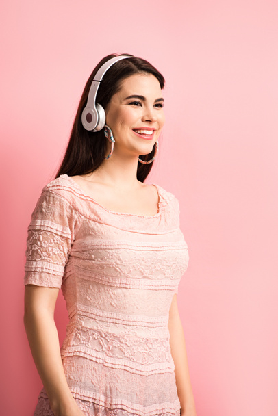 веселая девушка слушает музыку в беспроводных наушниках и смотрит в сторону на розовый фон
 - Фото, изображение