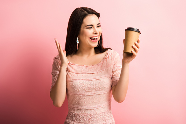 happy girl showing wow gesture while holding coffee to go on pink background - 写真・画像