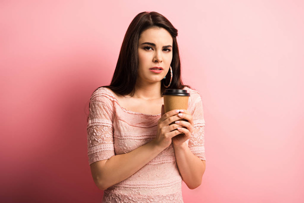 beledigd meisje houden koffie te gaan terwijl het kijken naar camera op roze achtergrond - Foto, afbeelding